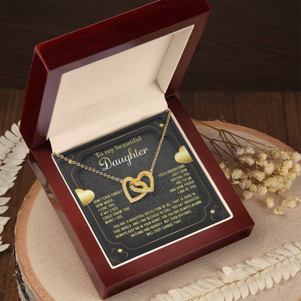 An open wooden jewelry box displays the "To Daughter, Beautiful Reflection - Interlocking Hearts Necklace" in an 18k yellow gold finish, adorned with delicate cubic zirconia crystals. The box also contains a heartfelt message addressed "To my beautiful Daughter" on a black card with white and yellow text, all set on a wooden surface with dried flowers.