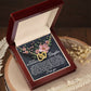 A wooden box holds the Daughter-Love Sight - Interlocking Hearts Necklace, featuring a gold heart pendant with pink flower accents. Inside the lid, a sentimental message is displayed in white text. The scene is completed by dried flowers near the box on the wooden table.
