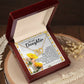 An open box reveals the Daughter-Journey Home - Interlocking Hearts Necklace, adorned with shimmering cubic zirconia crystals. The lid features a heartfelt message to a daughter, surrounded by yellow flower illustrations, resting on a wooden surface beside dried flowers, capturing beauty and emotion.