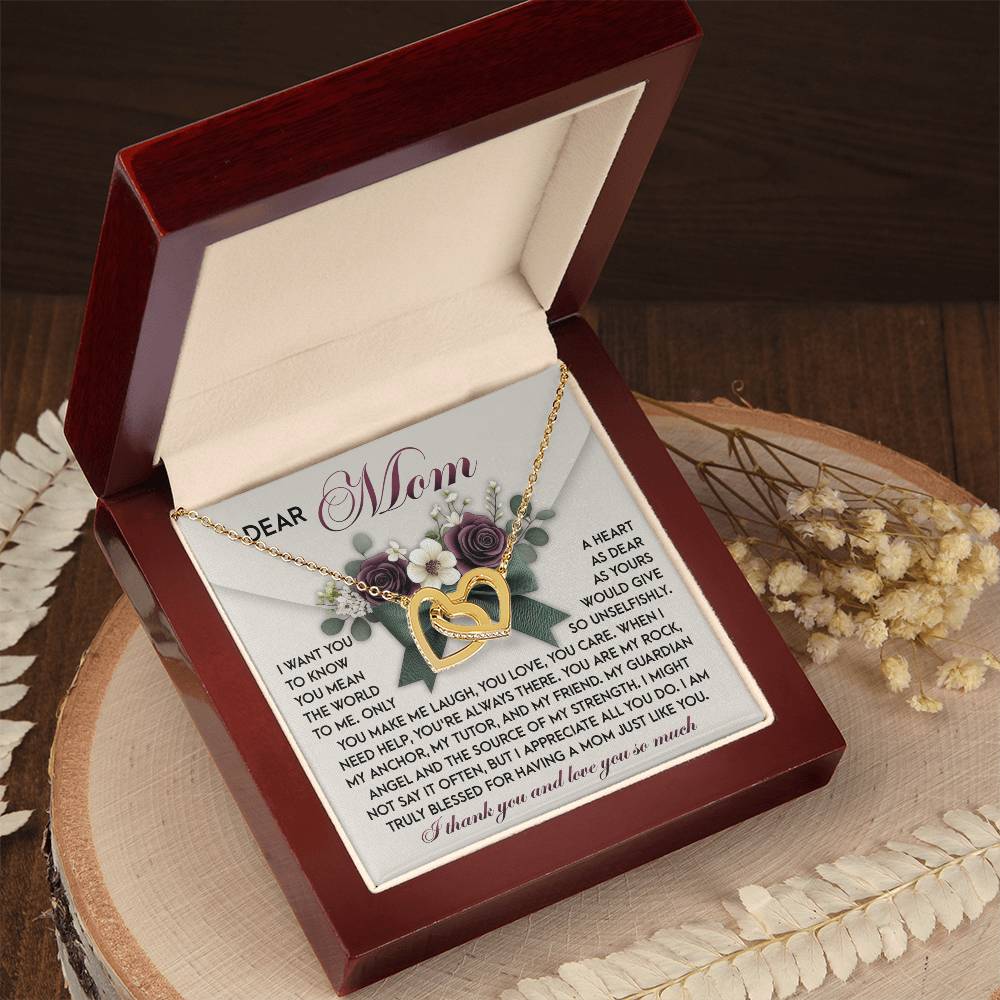An open jewelry box reveals the stunning "Mom-Guardian Angel - Interlocking Hearts Necklace" adorned with cubic zirconia crystals. Inside is a heartfelt poem for "Mom." The elegant box sits on a wooden surface, surrounded by dried flowers.