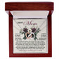 The Mom-Guardian Angel - Interlocking Hearts Necklace, embellished with sparkling cubic zirconia crystals, is elegantly presented in an open wooden box. Behind it, a heartfelt "Dear Mom" letter surrounded by floral designs conveys gratitude and admiration.