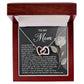 The Mom-Guiding Light Interlocking Hearts Necklace, with rose gold finish, is elegantly displayed in a wooden box. Its lid features a heartfelt message for "Mom" and a white rose illustration, while the soft interior lining gently cradles the exquisite piece.