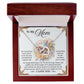 The Mom-Taught Me Interlocking Hearts Necklace, featuring a heart-shaped pendant with "MOM" in the center, colorful flowers, and cubic zirconia crystals, is presented in a red box. A heartfelt message behind it expresses gratitude and love for Mom’s support and lessons.