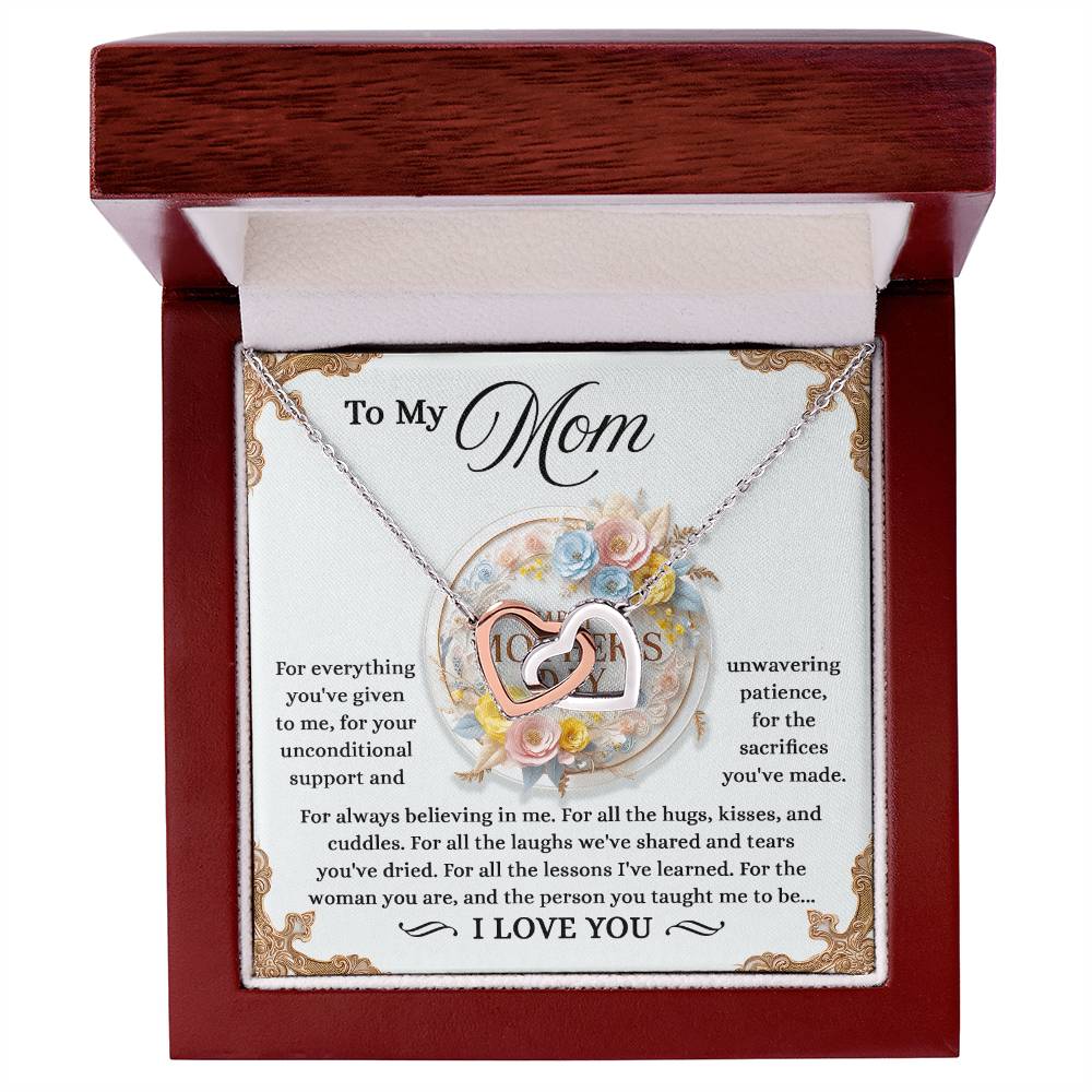 The Mom-Taught Me Interlocking Hearts Necklace, featuring a heart-shaped pendant with "MOM" in the center, colorful flowers, and cubic zirconia crystals, is presented in a red box. A heartfelt message behind it expresses gratitude and love for Mom’s support and lessons.