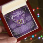 A hand holds an open jewelry box showcasing the Interlocking Hearts Necklace adorned with cubic zirconia, set against a purple butterfly-themed "To My Daughter" card with a heartfelt message. Blurred colorful lights create a warm backdrop.