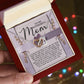 A person holds an open red jewelry box with the stunning Mom-Rare One - Interlocking Hearts Necklace made from surgical steel and adorned with cubic zirconia crystals, nestled against a heartfelt "Mom" card. In the background, colorful lights gently twinkle.