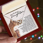 A hand holds a Daughter-Never Alone - Interlocking Hearts Necklace adorned with cubic zirconia, in a jewelry box featuring a heartfelt message and an illustration of a mother cat with her kitten. Softly twinkling colorful lights illuminate the scene.