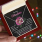 A hand presents an open jewelry box with a rose gold finish Interlocking Hearts necklace. The lid's message says, "To my Grandma, your love made me strong. Years of support and wisdom catch me when I fall. I love you forever." Colorful lights sparkle in the background.