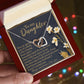 An open jewelry box held in hand reveals the Daughter-One Kind - Interlocking Hearts Necklace, crafted from surgical steel with a rose gold finish. Inside the lid, a heartfelt floral-accented message to a daughter is visible. Blurred multicolored lights in the background create a magical ambiance.