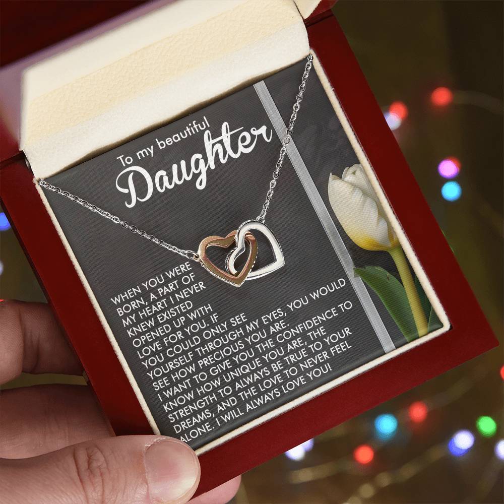 A hand holds an open jewelry box with a Daughter-Heart Love - Interlocking Hearts Necklace in rose gold. Inside, a heartfelt message for a daughter graces the card. Softly blurred colorful lights in the background add magic to the moment.