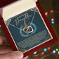 An open jewelry box held in a hand reveals the elegant Daughter-Little Princess - Interlocking Hearts Necklace, sparkling with cubic zirconia. A card inside says, "To my beautiful Daughter," with a heartfelt message. Colorful bokeh lights in the background enhance its enchantment.
