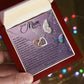 A hand opens a luxury box displaying the Mom-Shining Star - Interlocking Hearts necklace with cubic zirconia crystals. The lid shows a heartfelt message to "Mom" amid colorful butterflies and floral designs on a purple background, with softly blurred festive lights in the distance.