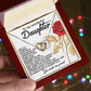A person holds an open jewelry box with the Daughter-Journey Challenges - Interlocking Hearts Necklace in rose gold. The lid has appreciative words for a daughter and a red rose illustration, against a softly blurred background with multicolored lights.