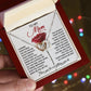 A hand holds an open red gift box with a card for a mom and the Mom-Be Fine - Interlocking Hearts Necklace featuring cubic zirconia crystals. The background is blurred with colorful bokeh lights, making it an ideal gift for her.