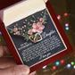 A person holds a Daughter-Love Sight box with an Interlocking Hearts necklace featuring pink flowers and cubic zirconia crystals. The lid has a loving message to a daughter, wishing her happiness. Blurred multicolored lights create a soft background ambiance.