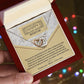 A hand holds an open red gift box revealing the Mother’s Day-Thank You - Interlocking Hearts Necklace with cubic zirconia crystals and a "Happy Mother's Day" card, with colorful blurred lights softly glowing in the background.