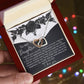 An open red jewelry box, held by hand, contains the Daughter-Better Person - Interlocking Hearts Necklace with cubic zirconia. A card inside features a heartfelt message to a daughter and floral designs. The background is softly blurred with multicolored lights.