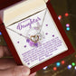 A hand holds an open jewelry box containing the Daughter-Angel Heart - Interlocking Hearts Necklace with cubic zirconia crystals. The box features a heartfelt message to a daughter from her mom, surrounded by purple flowers and heart decorations.