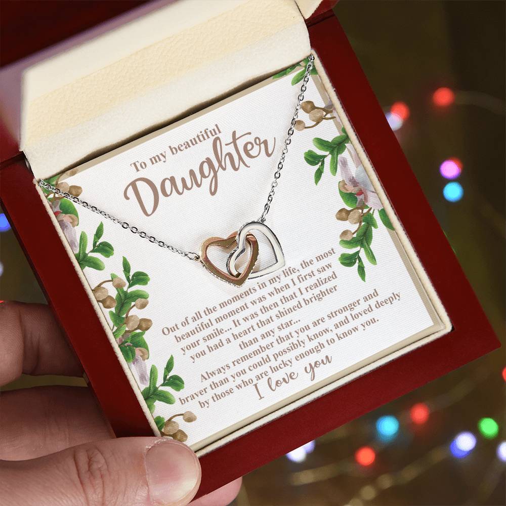 A hand holds an open red jewelry box revealing the Daughter-First Smile - Interlocking Hearts Necklace, crafted from lustrous surgical steel with a rose gold finish and sparkling cubic zirconia crystals. Inside, a card expresses love and pride for a daughter against a backdrop of blurred colorful lights.