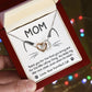 A hand holds an open jewelry box displaying the "Cat Mom - When I'm Mean" Interlocking Hearts Necklace with cubic zirconia crystals. The card features a cat face, reads "Mom," and includes a thank you from a "PurrFect Cat." Colorful lights enhance its rose gold finish's charm.