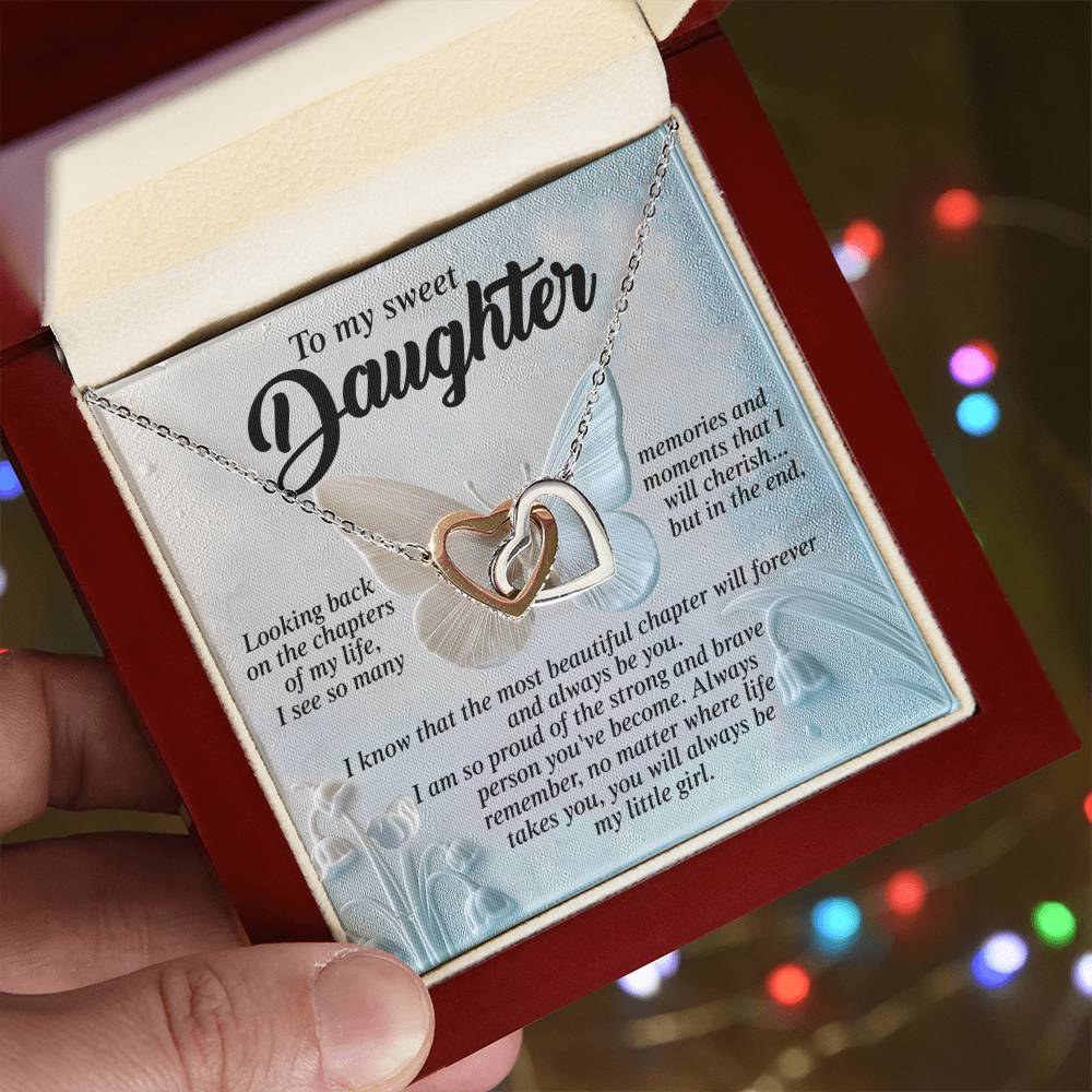 A hand opens a jewelry box to reveal the "Daughter-Beautiful Chapter - Interlocking Hearts Necklace" adorned with cubic zirconia crystals. The box's backdrop showcases a heartfelt message, as soft lights enhance the rose gold finish of the necklace.