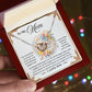 Someone holds a gift box containing a "Mom-Taught Me - Interlocking Hearts Necklace" with sparkling cubic zirconia crystals. The lid displays a heartfelt message for a mother, framed by floral designs, as colorful lights softly glow in the background.