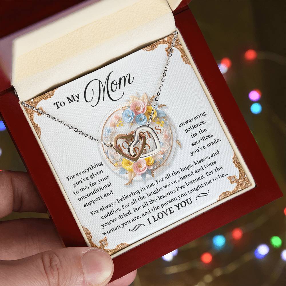 Someone holds a gift box containing a "Mom-Taught Me - Interlocking Hearts Necklace" with sparkling cubic zirconia crystals. The lid displays a heartfelt message for a mother, framed by floral designs, as colorful lights softly glow in the background.