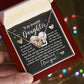 A hand holds an open red velvet box displaying the Daughter-Your Strength - Interlocking Hearts Necklace with a rose gold finish on a card reading, "To my beautiful daughter" with a supportive message. Blurred colorful lights twinkle warmly in the background, enhancing this heartfelt gift's elegance.