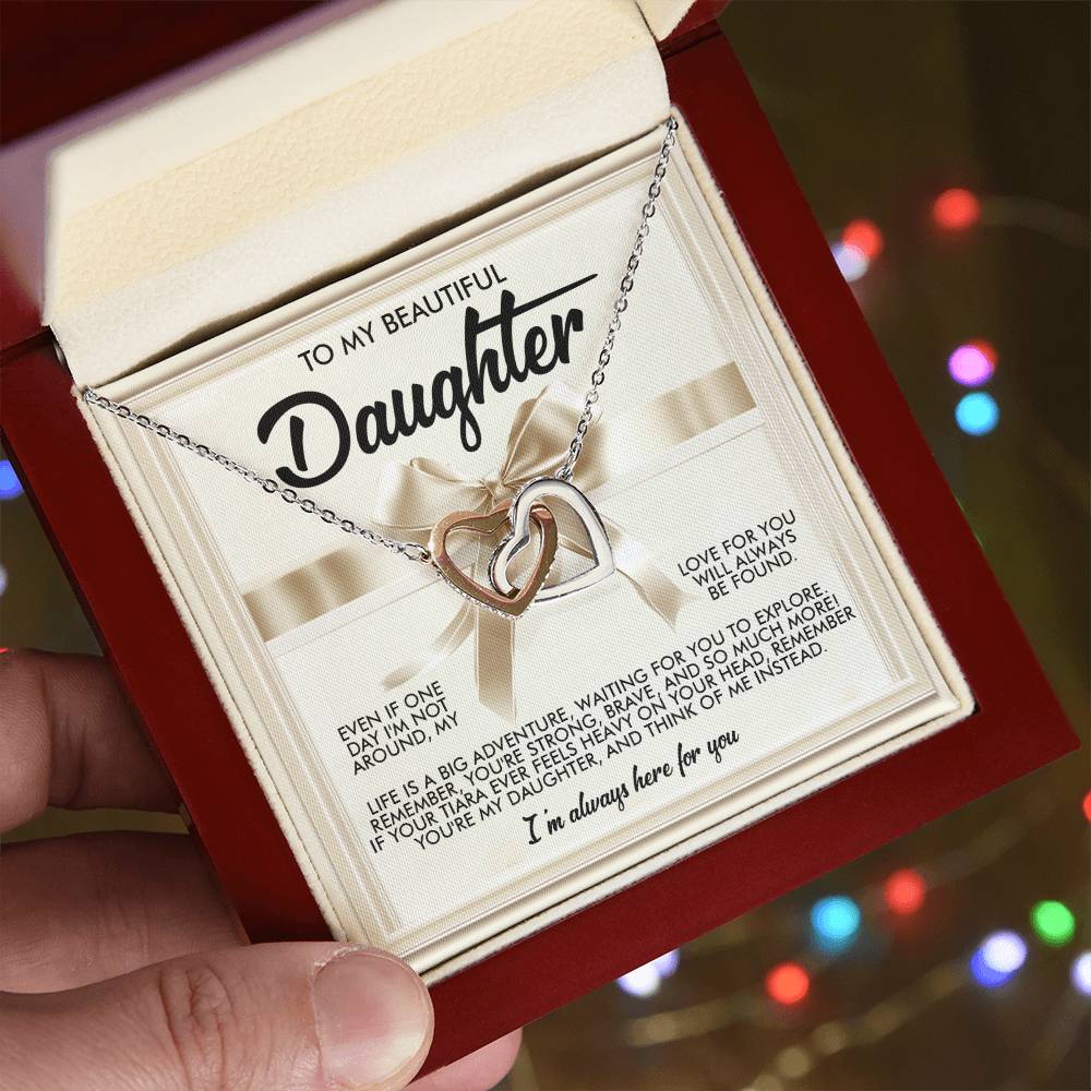 A hand presents an open red box containing the Daughter-Heavy Tiara - Interlocking Hearts Necklace. The silver piece shines with cubic zirconia crystals. Blurred colorful lights fill the background, and a heartfelt message for a daughter is elegantly shown on a card with decorative ribbon.