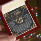 A hand holds an open red jewelry box that unveils the "Mom-God's Masterpiece - Interlocking Hearts Necklace," featuring heart-shaped pendants. The lid displays a heartfelt message about mothers as God's masterpiece, accentuated by rose gold and cubic zirconia crystals.