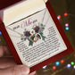 A person holds an open gift box featuring the "Mom-Guardian Angel - Interlocking Hearts Necklace" in rose gold, adorned with cubic zirconia crystals. A heartfelt message for "Mom" conveys gratitude and love, set against a softly lit background with colorful lights.