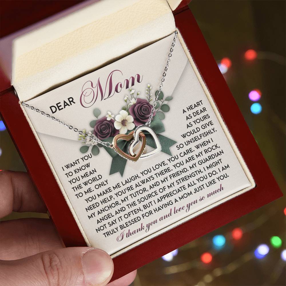 A person holds an open gift box featuring the "Mom-Guardian Angel - Interlocking Hearts Necklace" in rose gold, adorned with cubic zirconia crystals. A heartfelt message for "Mom" conveys gratitude and love, set against a softly lit background with colorful lights.