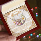 Someone holds an open jewelry box displaying the Daughter-Heart Happiness - Interlocking Hearts Necklace with cubic zirconia crystals over a floral card that says, "To my Daughter," against a blurred background of colorful lights.