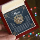 A hand holds an open jewelry box with the Mom-Thankful Heart - Interlocking Hearts Necklace, rose gold and cubic zirconia adorned. In the background, a heartfelt note to a boyfriend's mother expresses gratitude and admiration amid softly blurred twinkling lights.
