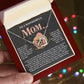 A hand holds an open gift box with a Mom-More To Me 2 - Interlocking Hearts Necklace adorned with cubic zirconia, with the lid featuring a sentimental message for Mom. Blurred colorful lights in the background make it perfect for an anniversary gift.