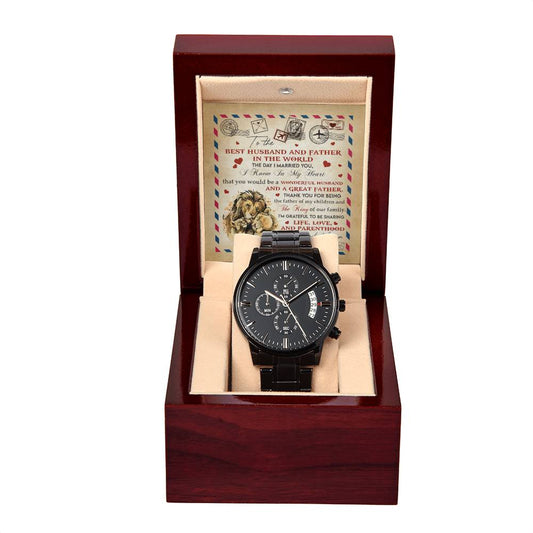 A black To Husband, Great Father - Metal Chronograph Watch with a copper dial is displayed in an open wooden box lined with beige fabric. Behind the watch, an illustrated card with text is positioned upright within the box.