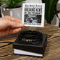 A hand holding a note designed like a newspaper announcing pregnancy above a box containing the To Father, The Baby Times - Love You Forever Bracelet—black braided with metallic accents, making the perfect gift for him to remember this special moment.