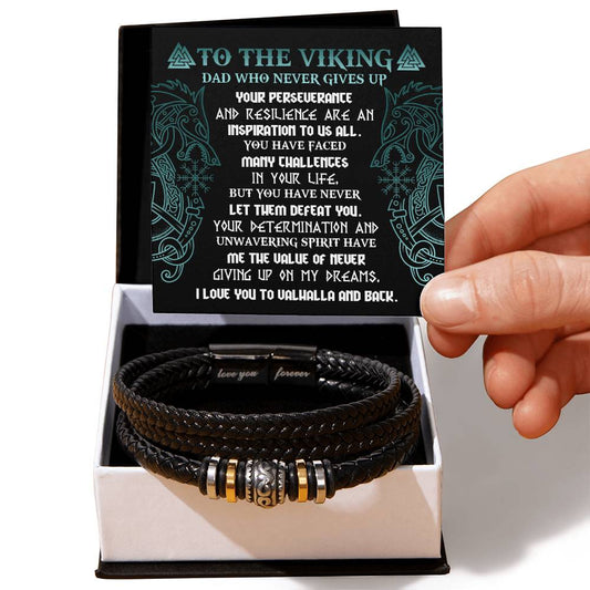 A hand holding a card with an inspirational message addressed "To Dad, Never Giving Up - Love You Forever Bracelet" above a black box containing a braided vegan leather bracelet with metal accents.