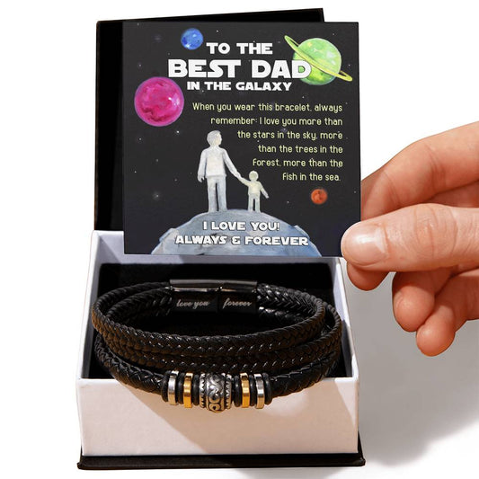 A hand holds a card that reads "To the Best Dad in the Galaxy" above a To Dad, In The Galaxy - Love You Forever Bracelet in a gift box. The bracelet is black with metallic accents, making it the perfect engraved gift for any special occasion.