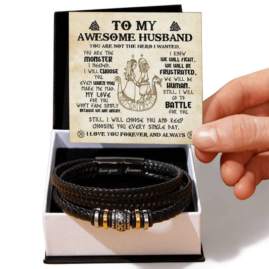 A person holding a note that reads "To My Awesome Husband" over a black braided vegan leather bracelet with metallic beads and symbols, placed in an open gift box labeled "To My Husband, Battle For You - Love You Forever Bracelet".