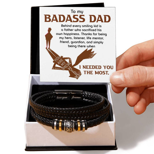 A hand holds a card that reads, "To my badass dad..." accompanied by a box containing a To Dad-, His Own Happiness - Love You Forever Bracelet crafted from black vegan leather with silver and gold accents.