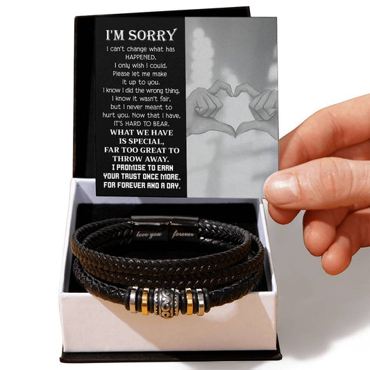 A hand holding an apology card over an open black box containing the "Sorry-Wish I Could - Love You Forever Bracelet," a black braided men's bracelet with metallic accents. The card features a heartfelt apology message, making this engraved bracelet a thoughtful gesture for any occasion.