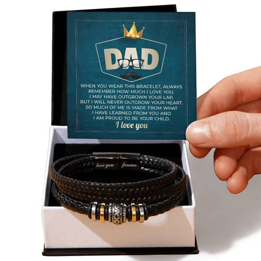 A hand holds a card with an engraved message for a dad over an open box containing a To Dad, Be Your Child - Love You Forever Bracelet made of black braided vegan leather with metallic accents.