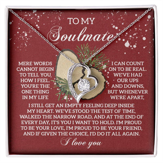 The Soulmate-Test Of Time - Forever Love Necklace, featuring a dazzling CZ crystal and a sophisticated gold finish, is elegantly presented in a box with a touching message to your soulmate on a red backdrop accompanied by pine decorations.