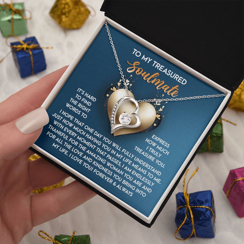 A hand holds an open jewelry box displaying the Soulmate-In My Life - Forever Love Necklace, featuring intertwined hearts with a sparkling CZ crystal and a white gold finish. The box cover reads "To My Treasured Soulmate," with a heartfelt message inside.