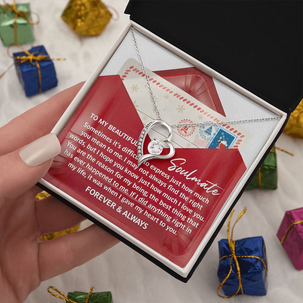 A hand holds an open gift box containing the Soulmate-Right Words - Forever Love Necklace, which is adorned with CZ crystals and comes with a card bearing the message "To my beautiful soulmate" along with a heartfelt note. The necklace's elegant white gold finish gleams as small wrapped presents are scattered in the background.