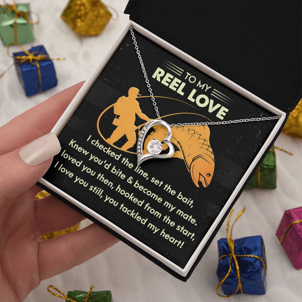 A hand holds an open black box showcasing the Soulmate-Tackled My Heart - Forever Love Necklace featuring a gold finish and CZ crystals. Beside it is a card with a fishing-themed romantic message, surrounded by small, colorful wrapped gifts in the background.