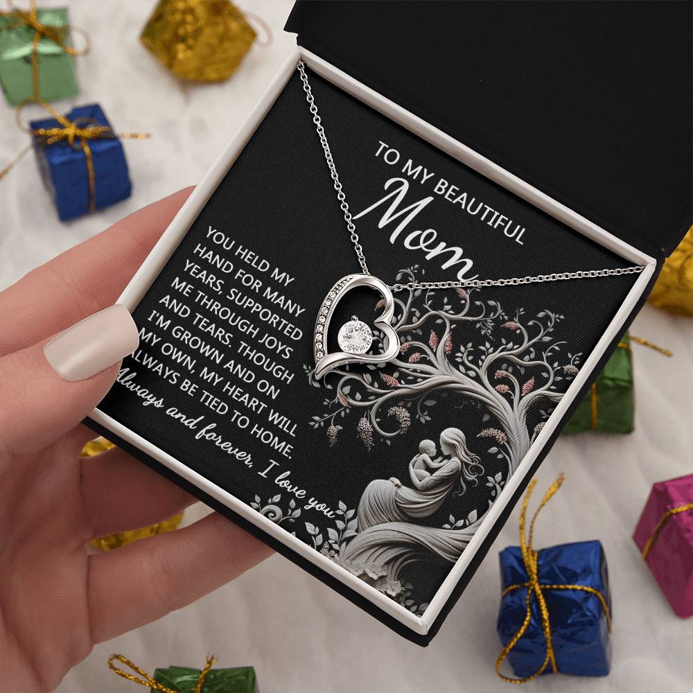 Hand holding an open black jewelry box with a "To Mom, On My Own - Forever Love Necklace" featuring a heart and two interlocked figures. Text inside the box reads a heartfelt message to "Mom." Small wrapped gifts are in the background.