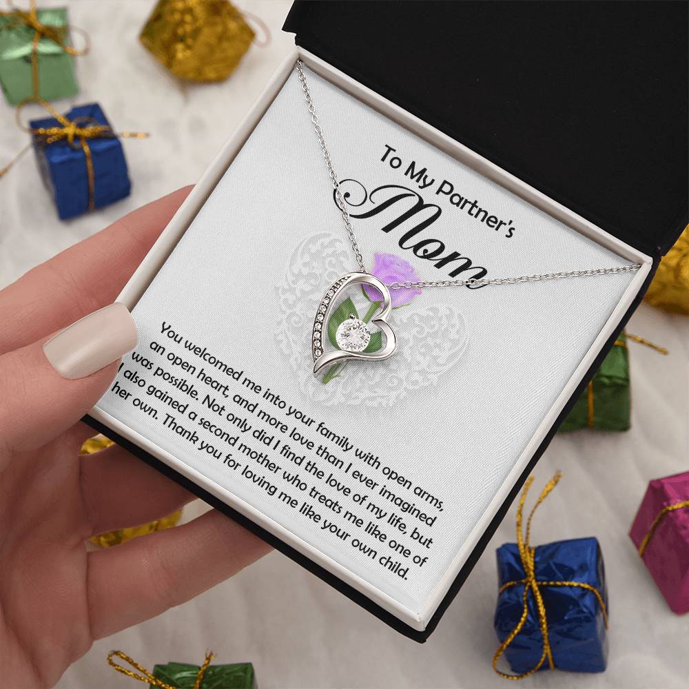 A hand holds an open box featuring the Partner's Mom-A Second Mother - Forever Love Necklace, a heart-shaped cubic zirconia piece with white gold finish and a loving message for "My Partner's Mom." Colorful gift boxes are scattered in the background.