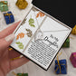 The Daughter-Biggest Fan - Forever Love Necklace, featuring a cubic zirconia stone and a radiant gold finish, is nestled in a heart-shaped gift box that conveys a parent's love to their daughter. Vibrant leaf patterns embellish the backdrop, and small gift boxes encircle this refined symbol of affection.